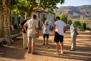Group of pétanque players discussing a shot on a shaded boulodrome with red earth, late afternoon light, and plane trees in Provence.